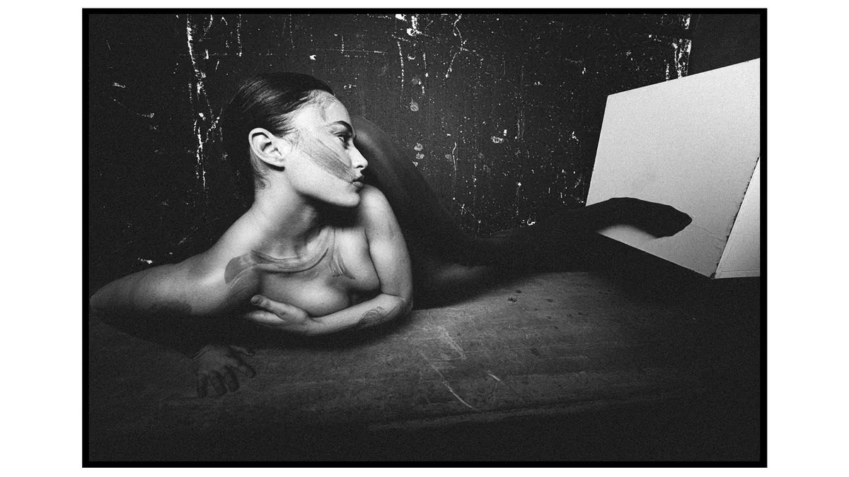 Reclining woman in profile with smears on her face and shoulder, in front of a dark scratched wall and a white panel