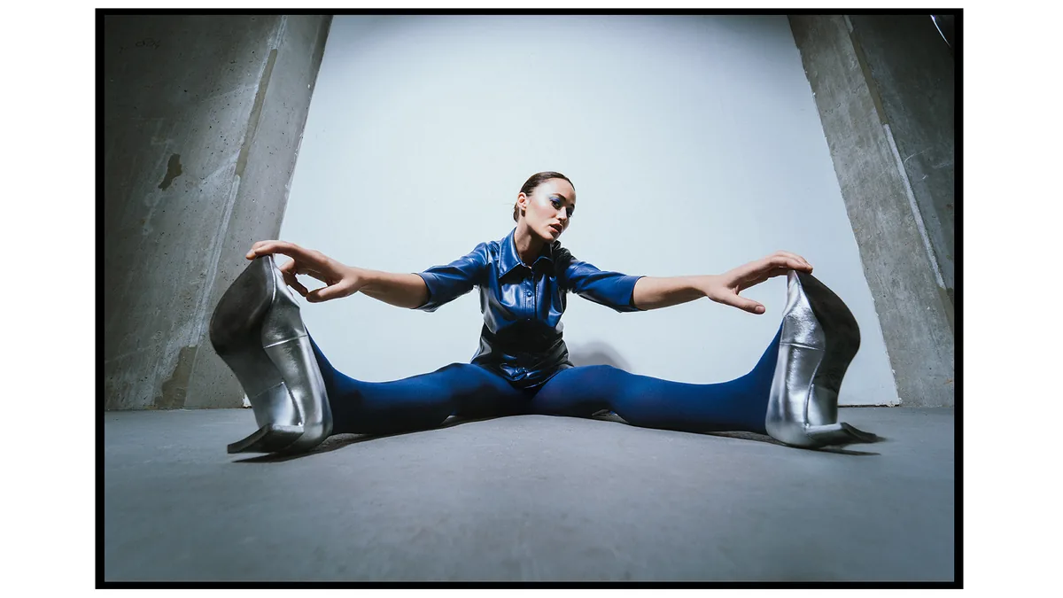 Woman in a blue leather shirt and silver heels sitting in a wide split, shot from a low angle between slanted concrete walls