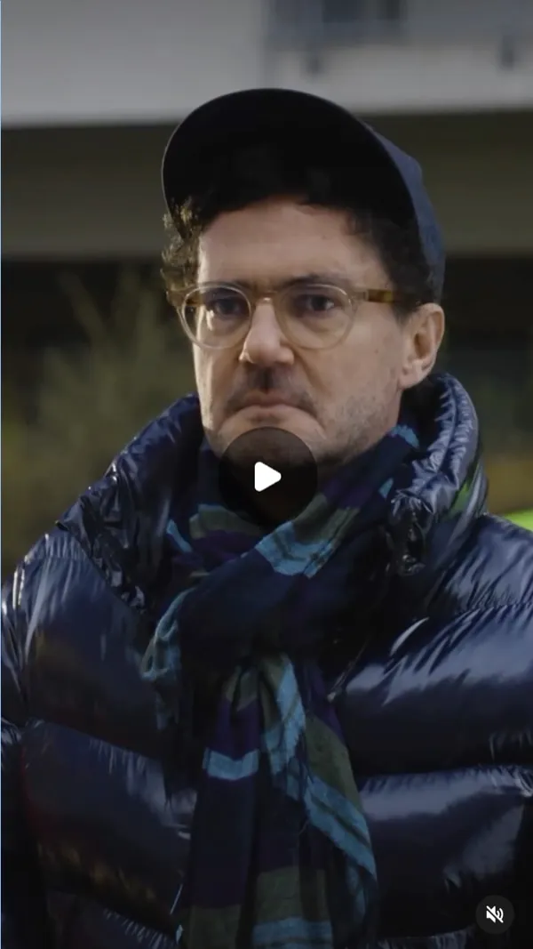 Reel: a serious man in glasses, a cap, a navy puffer jacket and a colourful scarf, blurred street behind