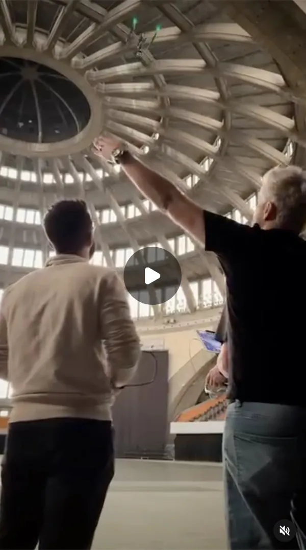 Reel: two men seen from behind in a hall with a ribbed dome, one raises an arm toward a small drone under the ceiling