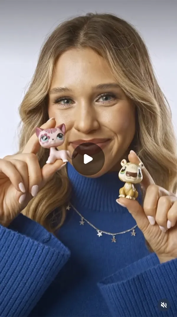 Reel: smiling woman in a blue sweater holds up two small animal figurines, grey backdrop