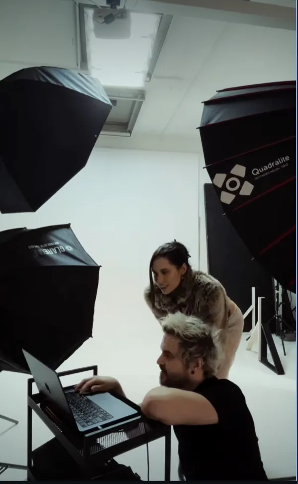 Behind-the-scenes reel: a woman in a furry jacket leans over a man at a laptop in a white studio with softboxes
