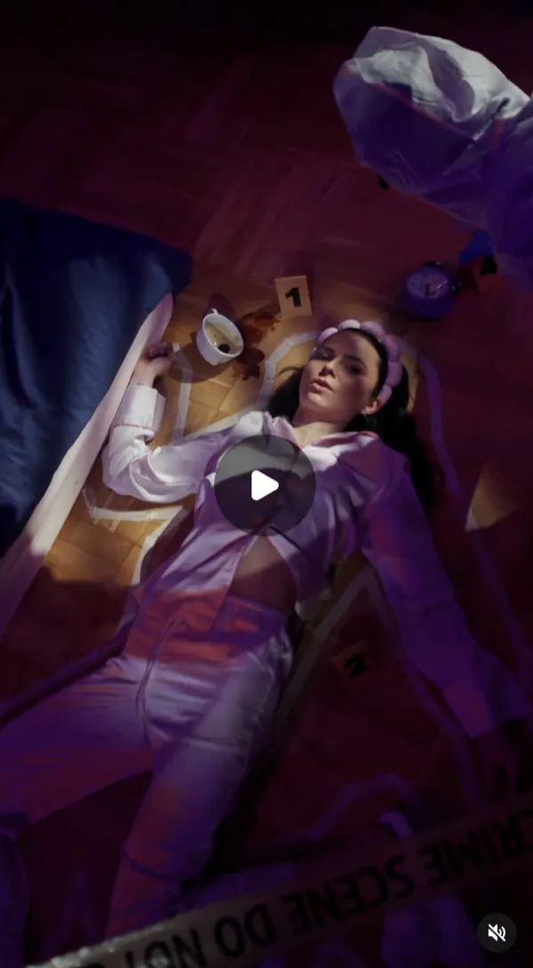 Reel: a woman in pink pyjamas lies on a parquet floor staged as a crime scene, chalk outline, markers and tape