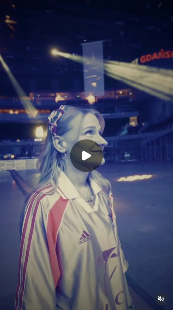 Reel: a woman in a light sports jacket and patterned scrunchie looks up in an empty arena, spotlight beams above