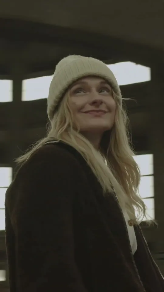 Short: smiling blonde woman in a cream beanie and dark fleece jacket looks up, hall windows behind her